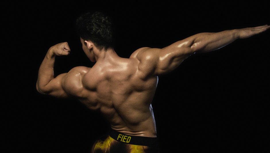 Back view of a male bodybuilder flexing muscles against a dark background.