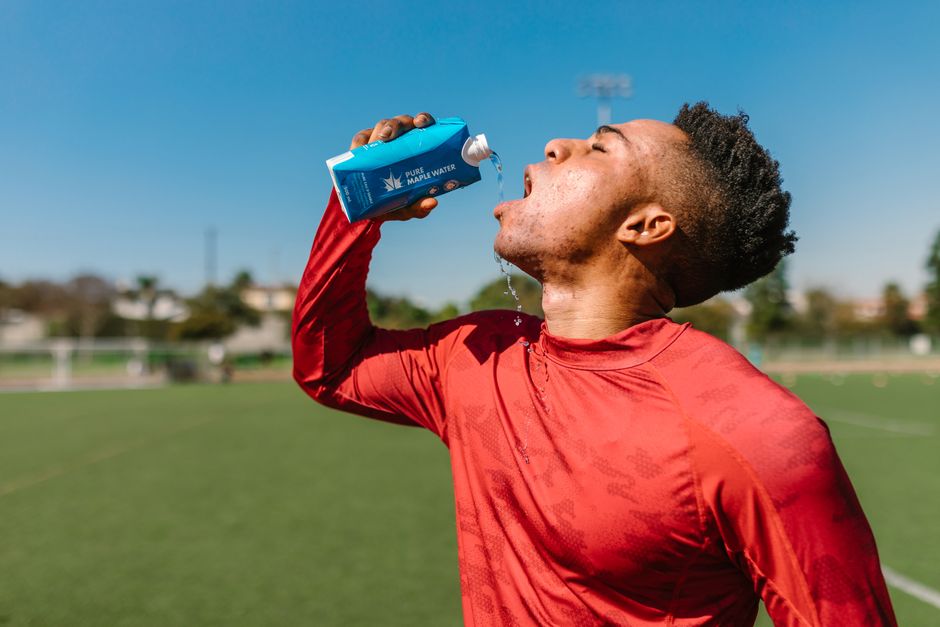 An athlete in a red shirt hydrates with maple water on a sunny day outdoors.