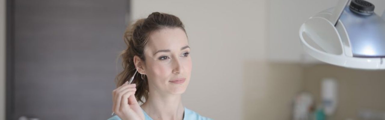 Positive young female doctor wearing blue uniform standing in light modern clinic with metal instruments in hand and looking away thoughtfully