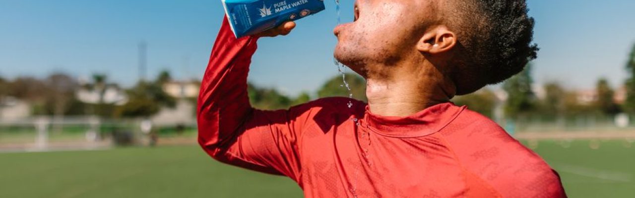 An athlete in a red shirt hydrates with maple water on a sunny day outdoors.